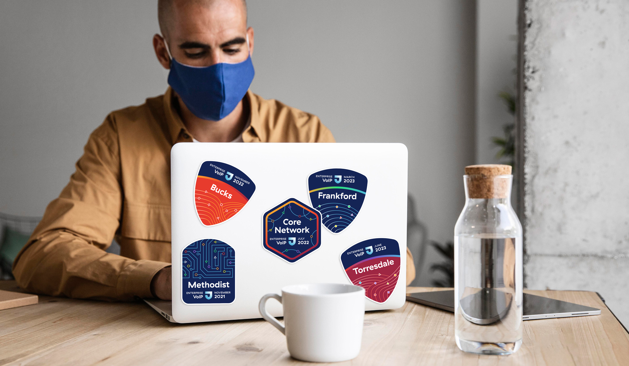 A man wearing a medical mask sits at a desk working on a laptop. The rear of the laptop is decorated with colorful stickers using the brand colors from Jefferson Health with a modern, technical illustration style.