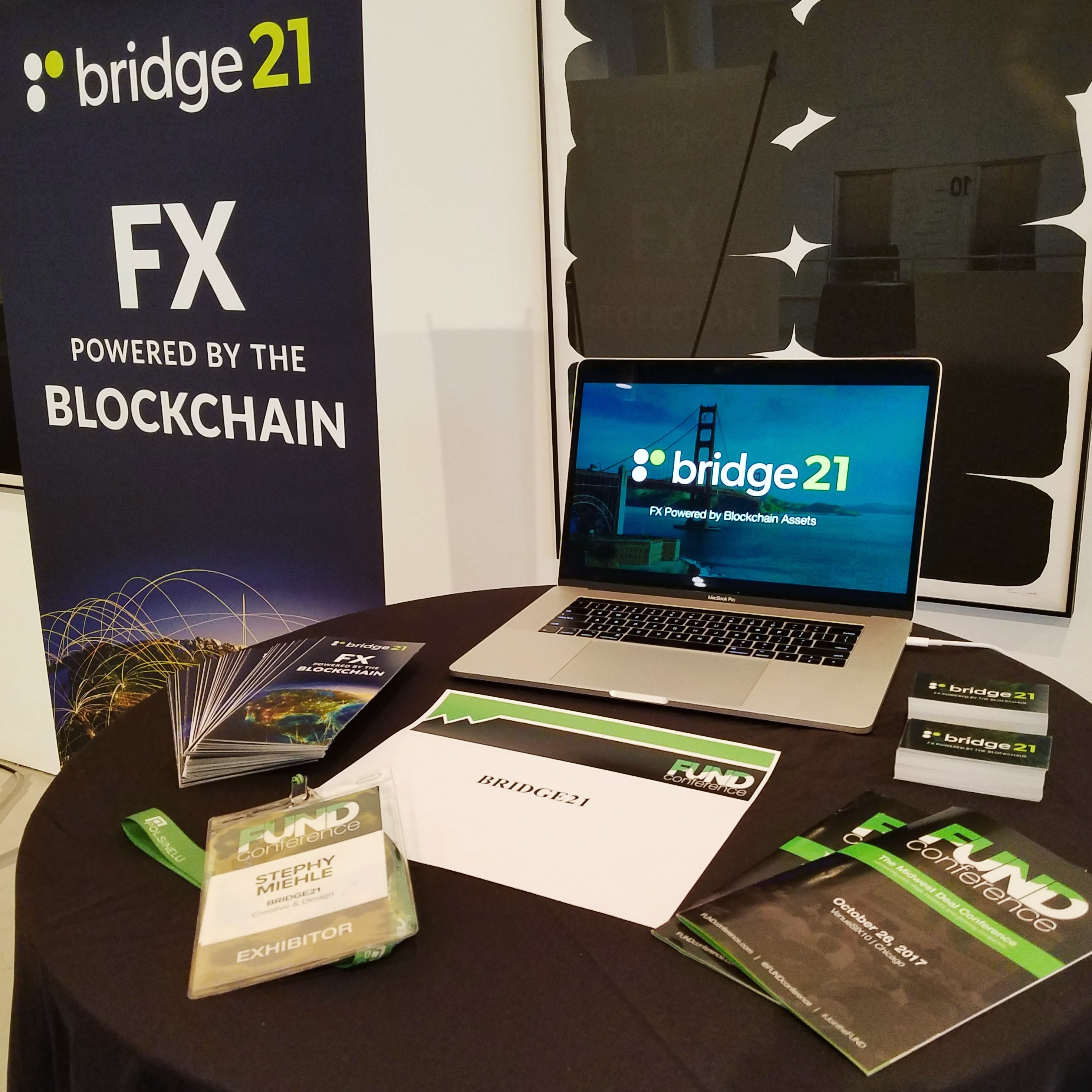 A table at the FUND Conference displaying information about bridge21's services, including a laptop with a demo video, brochures, and a large banner.