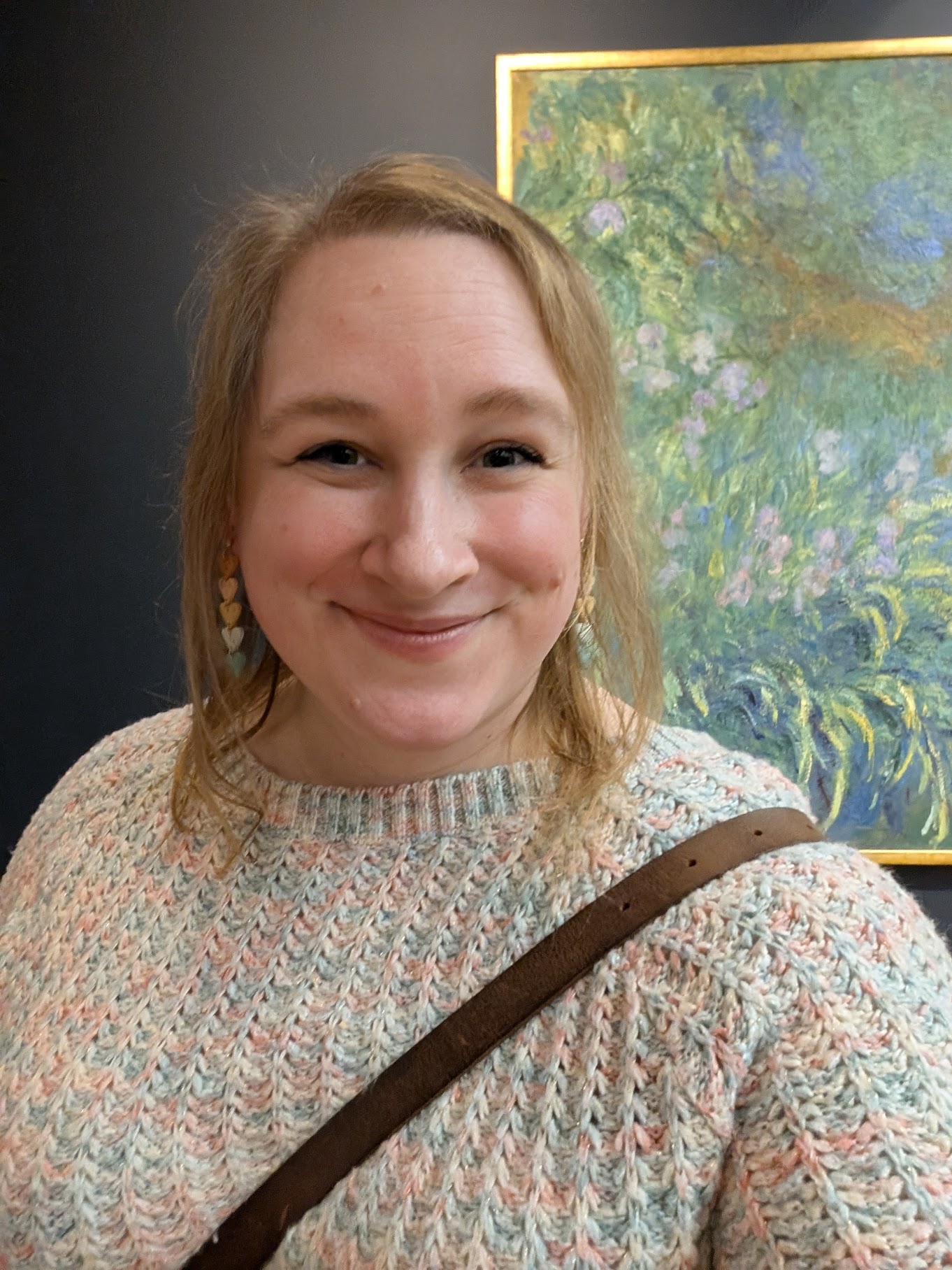 A close-up selfie of Stephy, a smiling, fair-skinned woman with shoulder-length, strawberry-blonde hair, standing indoors in the Art Institute of Chicago. She is wearing a chunky, multicolored knit sweater in soft shades of cream, pink, and blue. She has a dark brown leather strap from a bag across her right shoulder and heart-shaped cascading dangle earrings in pastel colors. The background is a dark gray wall, and over her left shoulder is the Impressionist Claude Monet painting titled 'Irises', featuring lush greens, yellows, wisteria purples, and pinks. The painting is in a golden frame.