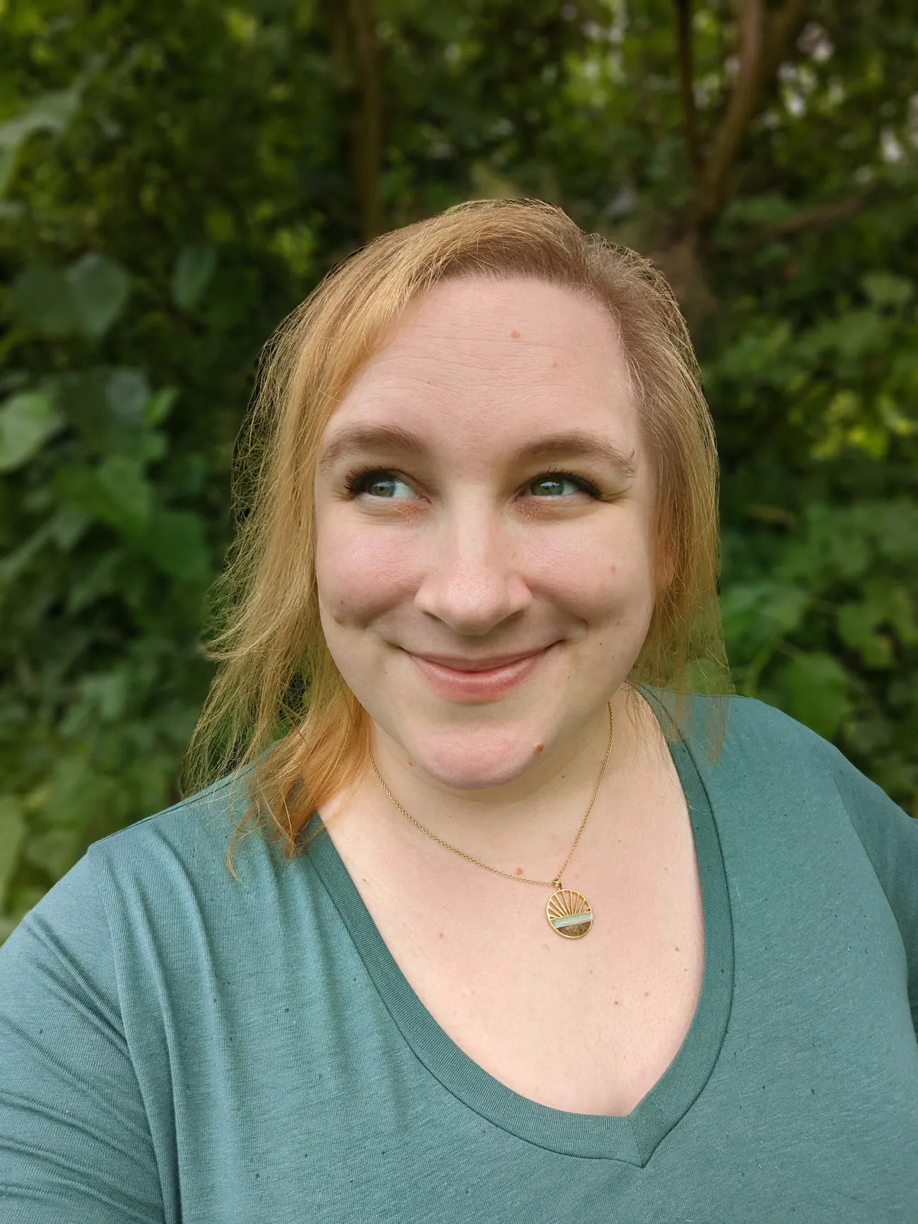 Stephy, a middle-aged white woman, stands outside in front of several trees. She has shoulder-length, windswept golden hair, green eyes, and a scattering of freckles and moles. She is wearing a casual blue-green V-neck shirt and a circular gold pendant necklace with a sunburst line motif above inlays of amazonite and dark brown sand. She is looking off into the distance with bright eyes and a slight smile.