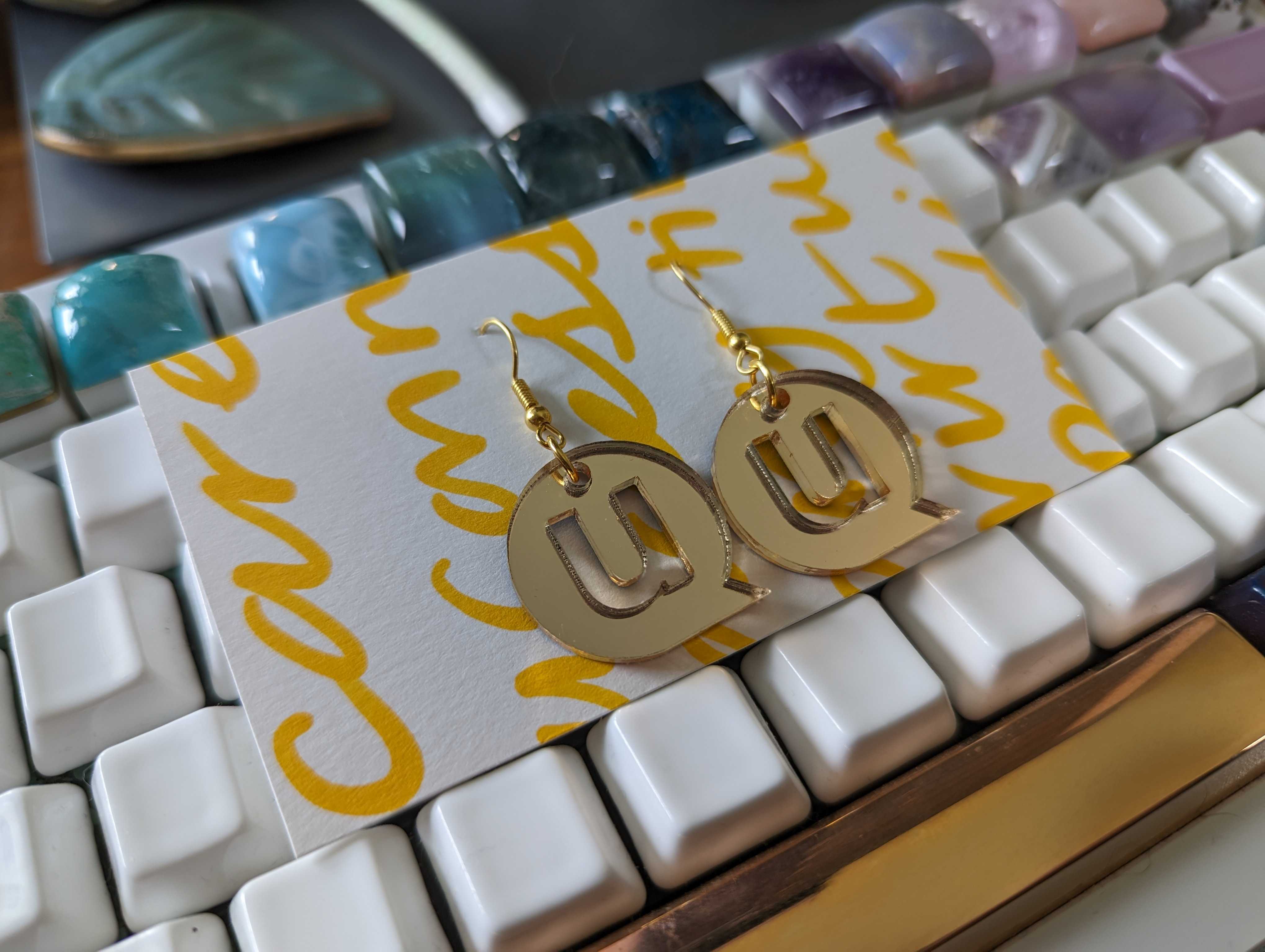 Metallic bronze acrylic earrings shaped like the uConnect logo. The earrings are displayed on a white card with bright yellow handwriting that creates visual texture. The card sits atop a computer keyboard.